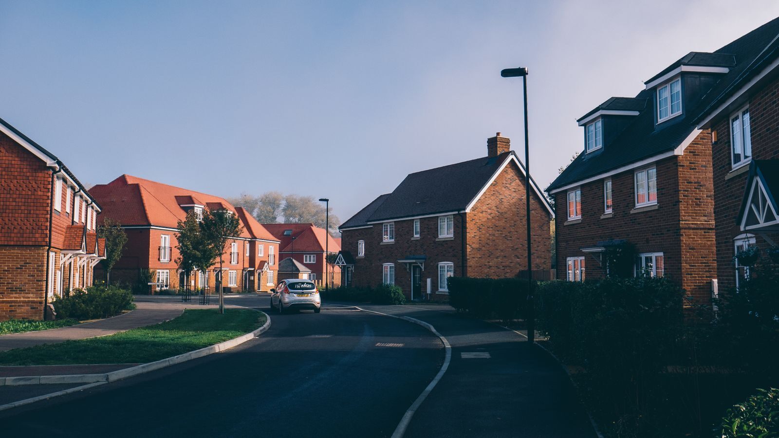 Residential street featuring modern brick houses with various roof styles, surrounded by greenery and a clear blue sky.