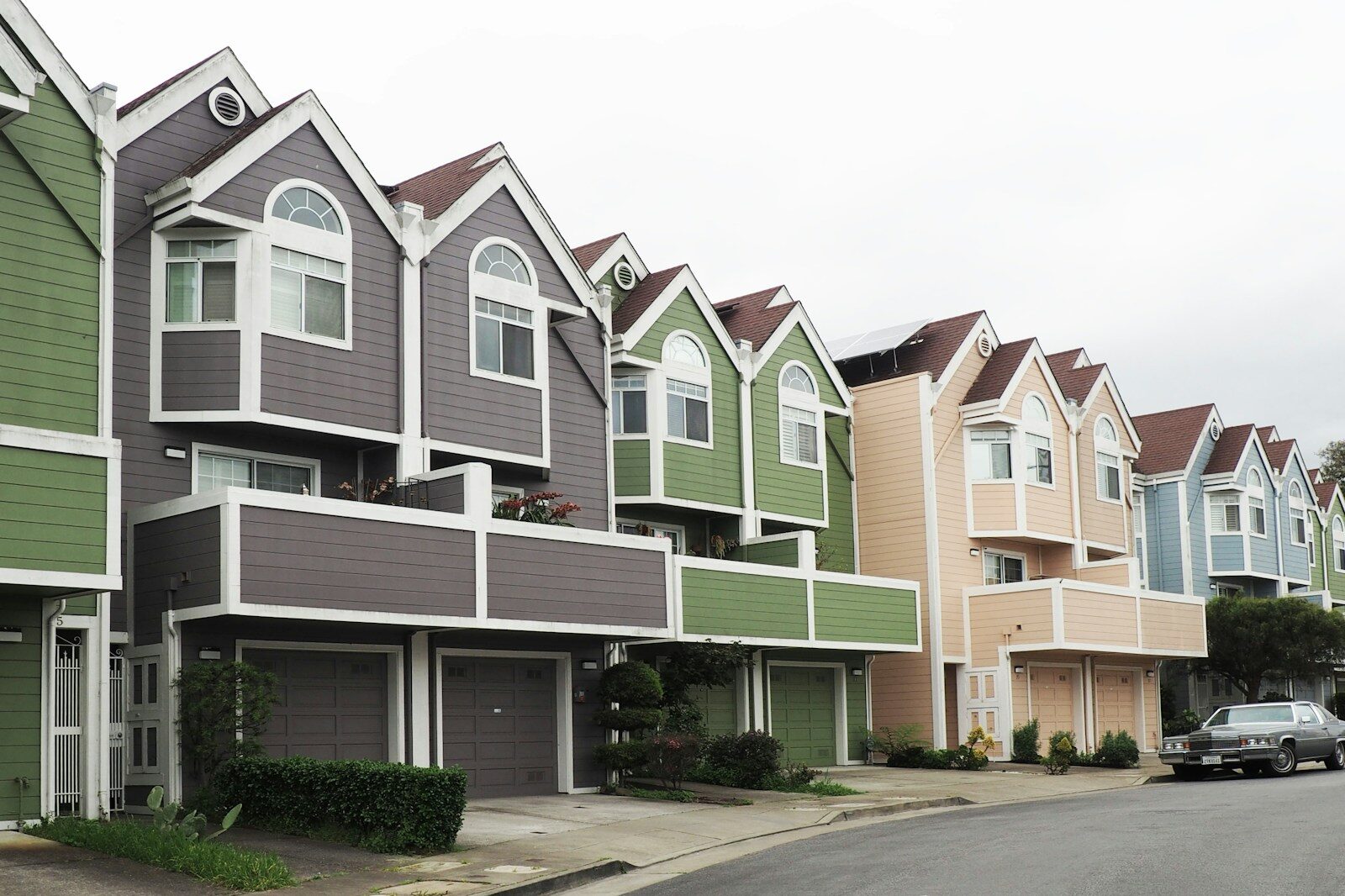 Colorful, modern townhouses with gabled roofs line a quiet street, showcasing a mix of green, gray, pink, and blue exteriors.