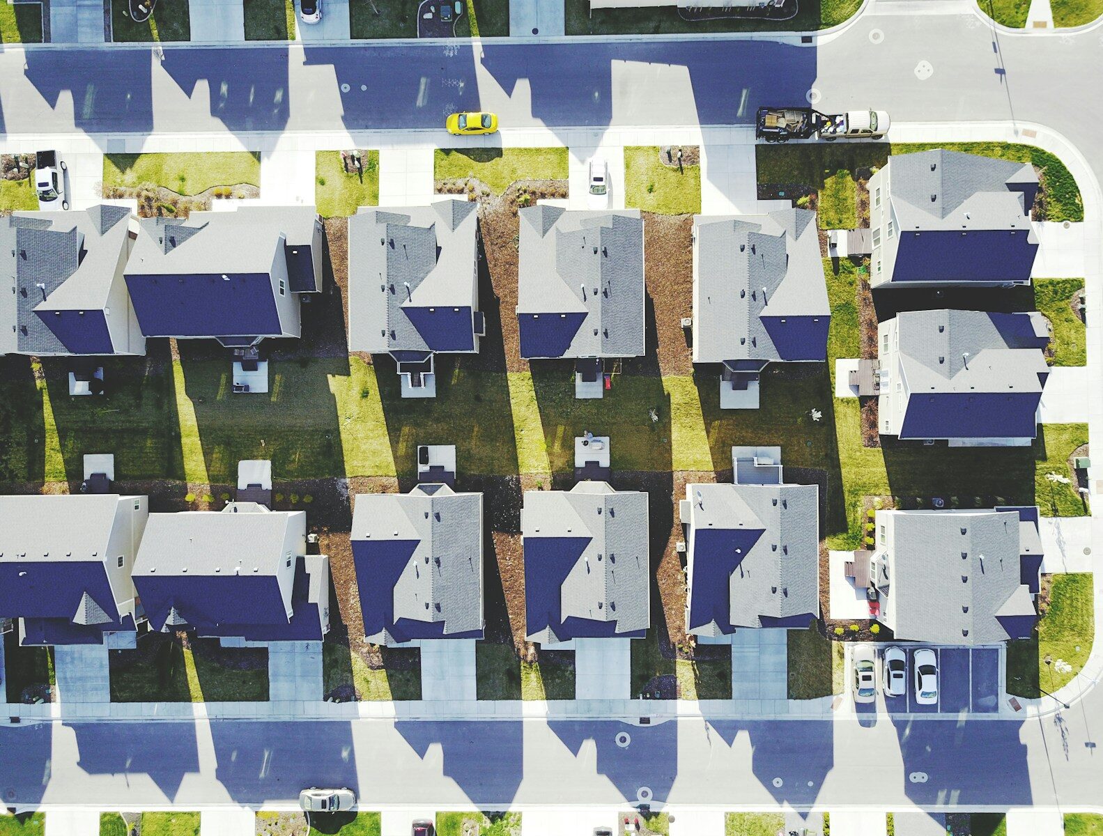 Aerial view of a suburban neighborhood showcasing rows of houses with shadows, green lawns, and parked cars on the streets.